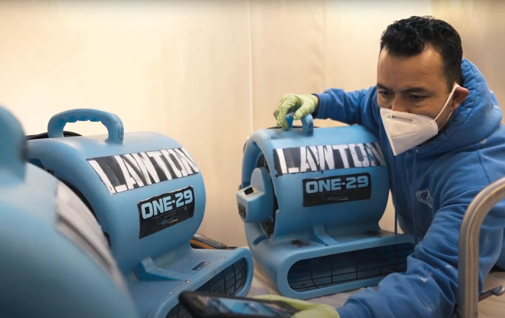 Professional water damage restoration technician using industrial water extraction equipment with blue air movers in damaged room
